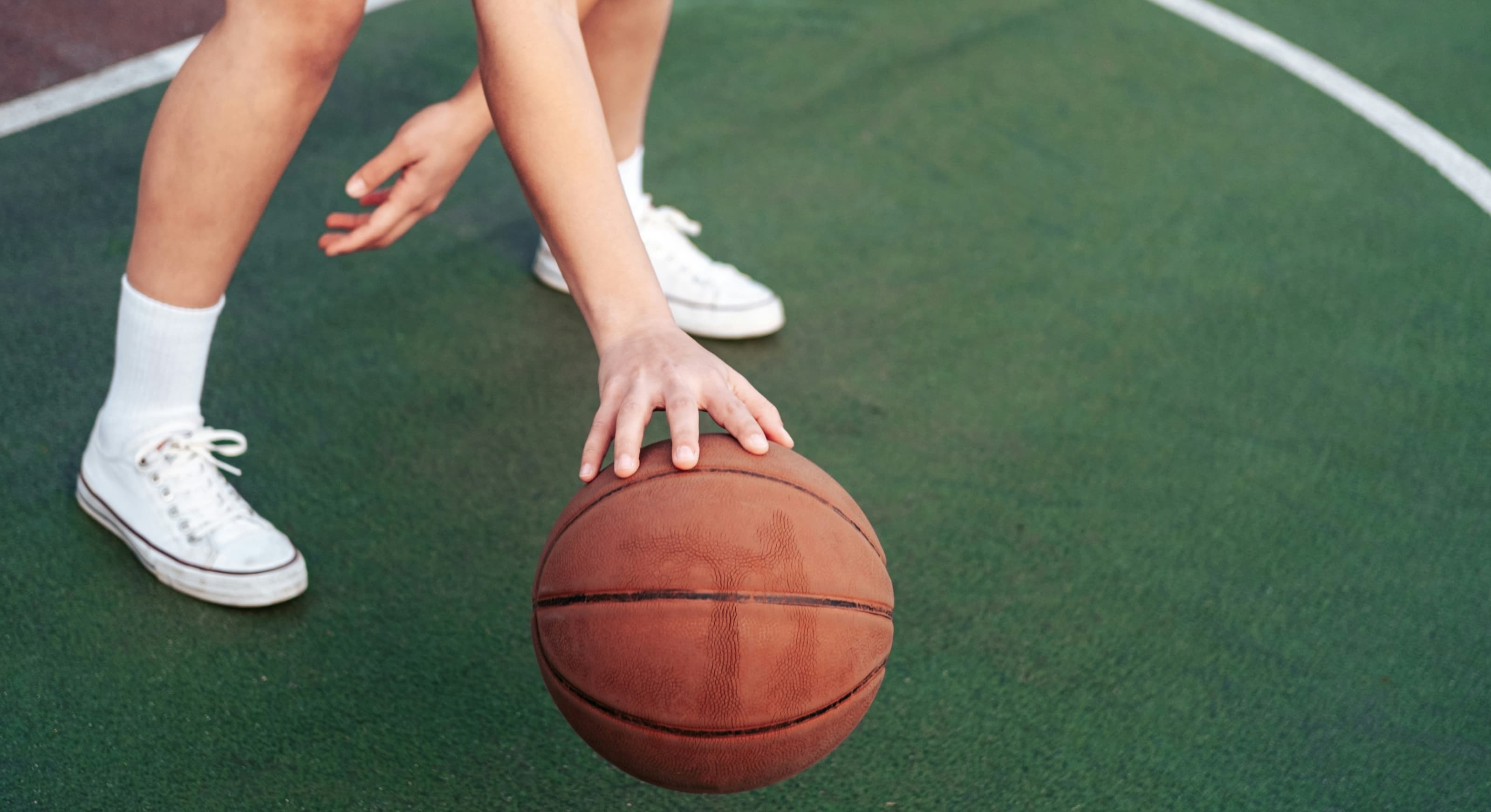 Person in white sneakers dribbling a basketball on a green outdoor court.